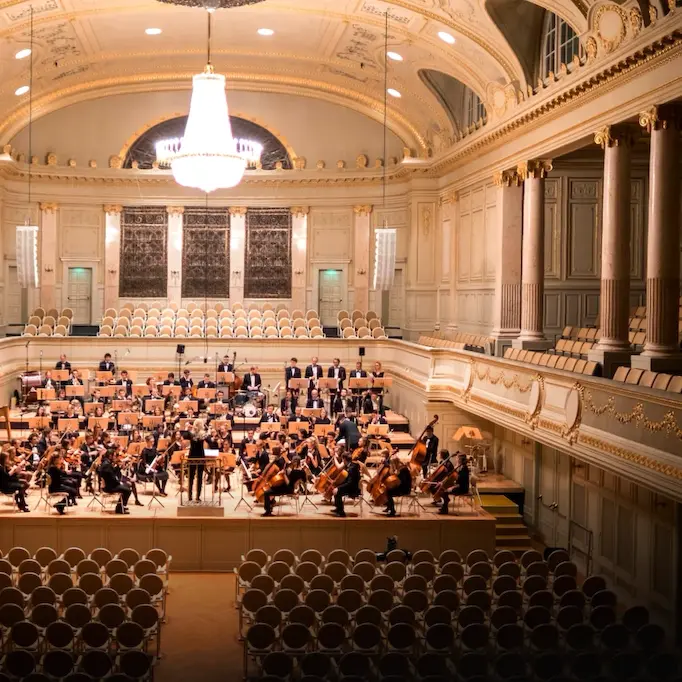 Orchestra rehearsing in an elegant concert hall with a large chandelier and ornate decor.