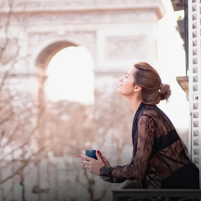 Woman enjoying a cup of coffee on a balcony with the Arc de Triomphe in the background.