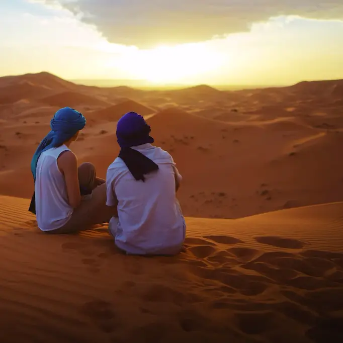 Two people in headscarves sitting on sand dunes watching the sunset in the Sahara Desert.