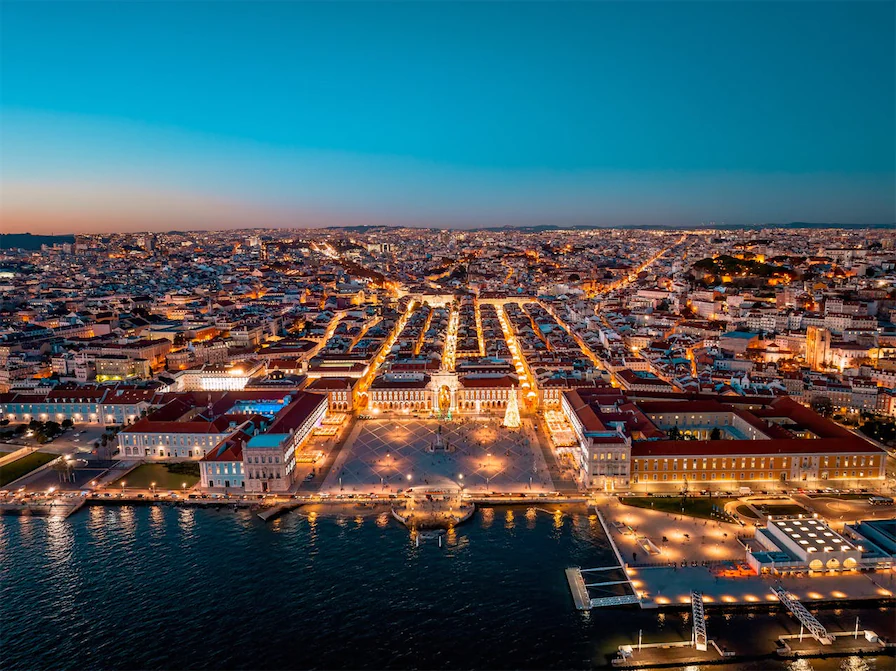 Aerial view of Lisbon at dusk, with the city lit up, highlighting its historic squares, buildings, and waterfront
